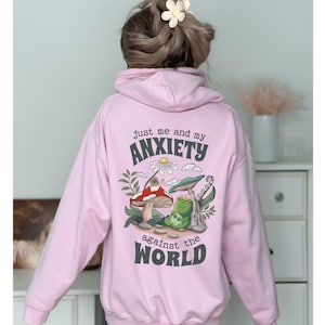 May include: Pink hoodie with a graphic print of a frog, mushroom, and snail with the text "Just me and my anxiety against the world".