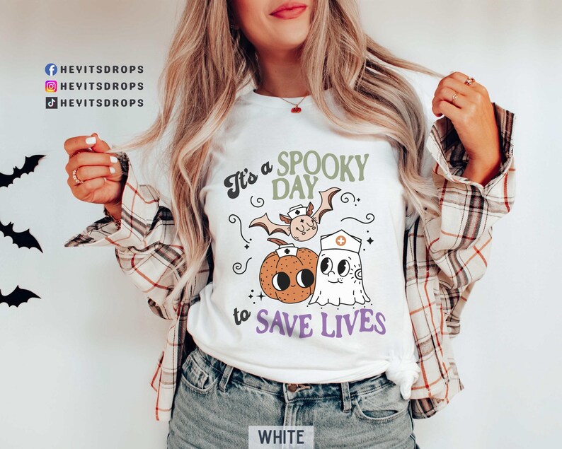 Puede incluir: Camiseta blanca con un dise&ntilde;o de Halloween. El dise&ntilde;o presenta una calabaza, un murci&eacute;lago y un fantasma con gorro de enfermera, con el texto "It's a Spooky Day to Save Lives". La camiseta se combina con una camisa a cuadros y vaqueros azules.
