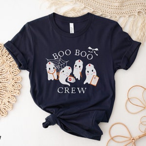 May include: Navy blue t-shirt featuring a white graphic of three ghosts dressed as nurses. The ghosts are holding medical supplies and the text "Boo Boo Crew" is printed on the shirt.