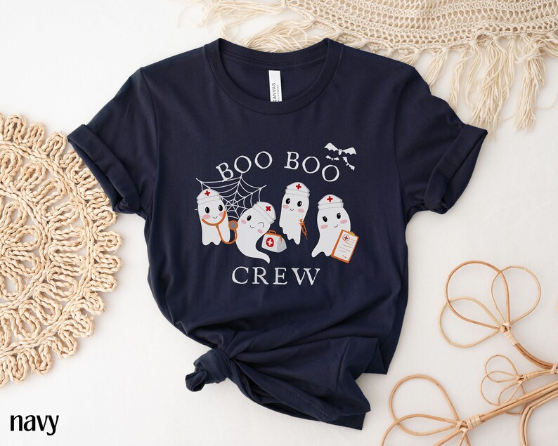 May include: Navy blue t-shirt featuring a white graphic of three ghosts dressed as nurses. The ghosts are holding medical supplies and the text "Boo Boo Crew" is printed on the shirt.
