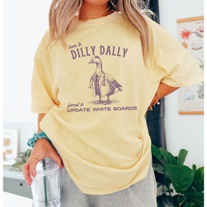 May include: Yellow t-shirt with a graphic of a duck wearing a doctor's coat and stethoscope. The text on the shirt reads "born to dilly dally forced to update white boards".
