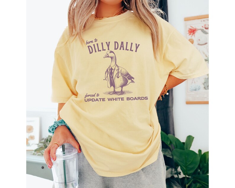May include: Yellow t-shirt with a graphic of a duck wearing a doctor's coat and stethoscope. The text on the shirt reads "born to dilly dally forced to update white boards".