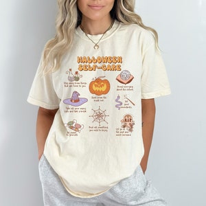 May include: A beige t-shirt with a graphic print of Halloween self-care tips. The graphic includes a pumpkin, a witch's hat, a spiderweb, a potion bottle, a wand, a graveyard, and candy. The text on the shirt reads "Halloween Self-Care".