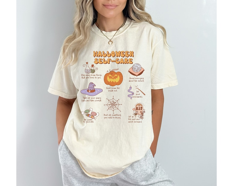 May include: A beige t-shirt with a graphic print of Halloween self-care tips. The graphic includes a pumpkin, a witch's hat, a spiderweb, a potion bottle, a wand, a graveyard, and candy. The text on the shirt reads "Halloween Self-Care".