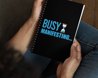 May include: A black spiral notebook with the text "BUSY MANIFESTING..." in blue and a blue hourglass graphic.