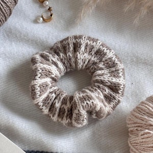 Può includere: Un primo piano di un elastico per capelli in maglia marrone e bianco su una coperta bianca e grigia. Ci sono due gomitoli di lana, uno marrone e uno rosa, e un paio di orecchini d'oro con accenti di perle. Un libro di cucina con il titolo "Festive Breads" è aperto sulla coperta.