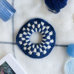 May include: A blue and white knitted hair scrunchie with a checkered pattern. The scrunchie is lying on a white and blue striped blanket.