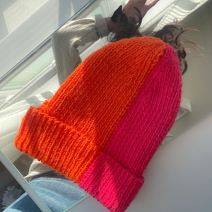 Puede incluir: Un gorro de punto con un dise&ntilde;o de bloques de color horizontales. La mitad superior del gorro es naranja y la mitad inferior es rosa.