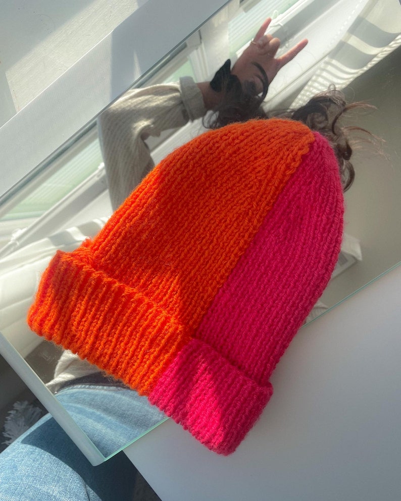 Puede incluir: Un gorro de punto con un dise&ntilde;o de bloques de color horizontales. La mitad superior del gorro es naranja y la mitad inferior es rosa.