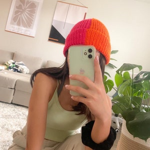 Puede incluir: Una persona lleva una boina de punto rosa y naranja con un top verde sin mangas y pantalones beige. La persona sostiene un iPhone verde claro en su mano.