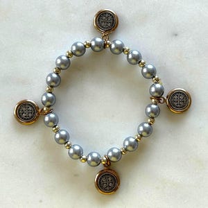 May include: Gray beaded bracelet with gold accents and four round charms. The charms have a detailed design in a gold frame. The bracelet is on a white marble surface.