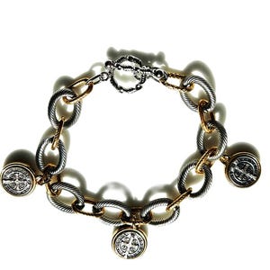 May include: A silver and gold-toned bracelet with a toggle clasp. The bracelet features oval-shaped links and gold accents. Three round charms with a religious emblem are attached to the bracelet.