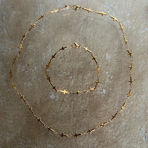 May include: A gold-toned metal jewelry set consisting of a necklace and a matching bracelet. Both pieces feature a delicate chain design with small, evenly spaced cross-shaped charms integrated throughout the length of each item, secured by clasps.
