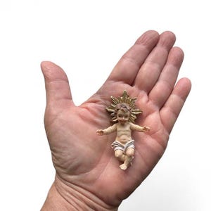 May include: A small figurine of the baby Jesus with a gold halo. The figure is depicted with outstretched arms and wearing a white cloth. The figurine is held in the palm of a hand against a white background.