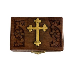 May include: A rectangular dark brown wooden box featuring a polished gold-tone metal cross centered on the lid. The lid is decorated with carved floral patterns on both the left and right sides of the cross, and a gold-tone metal latch is visible.