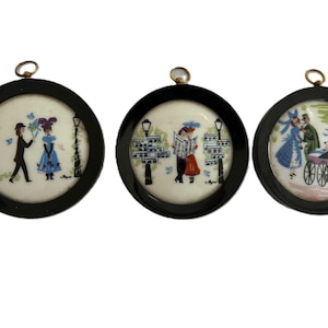May include: Three black framed circular porcelain ornaments with white backgrounds and colorful painted scenes of couples walking in a city setting.