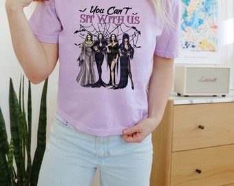 May include: A light purple t-shirt with the text "You Can't Sit With Us" and a graphic of four women in gothic attire. The design includes bats and a spiderweb. The shirt is paired with light blue jeans.