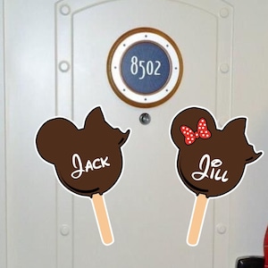 May include: Two chocolate Mickey Mouse head ice cream pops with sticks. The Mickey Mouse head on the left has the name "Jack" written on it. The Mickey Mouse head on the right has the name "Jill" written on it and a red bow with white polka dots. The pops are on a white door with a round porthole with the number "8502" on it.