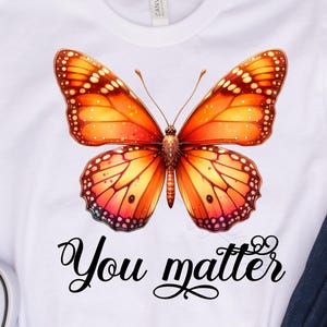 Può includere: T-shirt bianco con una farfalla arancione e gialla e la scritta "You matter" in nero. L'immagine include jeans blu e scarpe da ginnastica grigie.