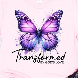 以下が含まれることがあります： 紫と青の色合いの水彩画の蝶のデザインが特徴の、明るいピンク色のTシャツ。「Transformed by God's Love」の文字が下にプリントされています。このシャツは、青いジーンズと白いスニーカーと組み合わせています。