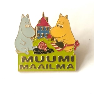 Op de afbeelding: Een kleurrijke emaille pin met twee stripfiguren, een lichtblauwe en een gele, met een rood en blauw gebouw op de achtergrond. De pin heeft de tekst "MUUMI MAAILMA" in groen en goud.
