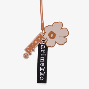Könnte beinhalten: Ein brauner und schwarzer Marimekko-Schlüsselanhänger mit einem Blumenanhänger. Der Anhänger ist eine hellrosa Blume mit brauner Mitte. Der Schlüsselanhänger hat den Text "Marimekko" in weiß aufgedruckt.