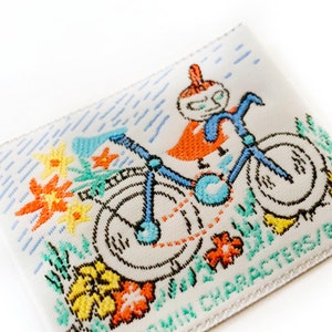 May include: A white woven label featuring a colourful illustration of a character riding a bicycle. The character is wearing a red and orange outfit and a blue helmet. The bicycle has blue wheels and a green frame. The label includes the text "MOOMIN CHARACTERS/BULLS" in green.
