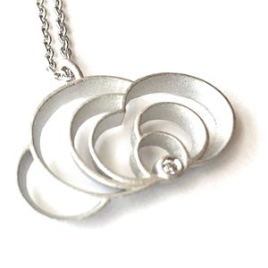 May include: A silver pendant necklace featuring a layered, openwork design of concentric circles. A small, clear gemstone is set in the center of the pendant. The necklace has a delicate silver chain and is set against a white background.