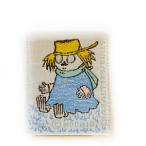 May include: A white fabric patch with a cartoon illustration of a girl wearing a yellow hat and blue dress. The text "MOOMIN" and "CHARACTER  SAABELS" is printed on the patch.