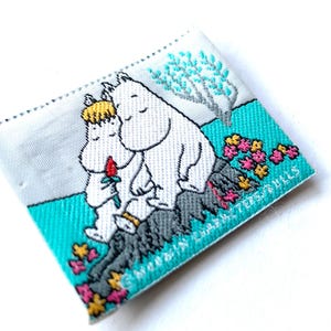 Puede incluir: Parche bordado con dos personajes de dibujos animados blancos abrazándose, con una flor roja. El parche tiene un borde turquesa con flores rosas y amarillas, una roca gris y un árbol. El texto dice "MOOMIN CHARACTERS DOLLS".