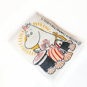 Puede incluir: Parche de tela blanco con una imagen de dibujos animados de un personaje de Moomin, una criatura blanca con una flor en la cabeza, sosteniendo un objeto a rayas rojas y amarillas. El texto "MOOMIN CHARACTER" está impreso en el parche.