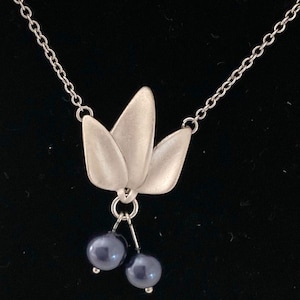 Puede incluir: Un collar de tono plateado con un colgante en forma de tres hojas lisas de acabado mate. Dos pequeñas cuentas esféricas de color azul oscuro cuelgan de la base del colgante de hojas, suspendidas por finos alambres metálicos plateados.