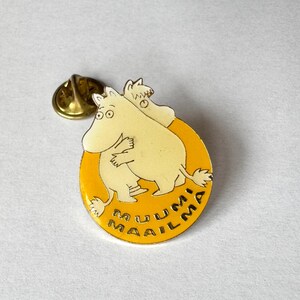 May include: A round metal enamel pin featuring two cream-colored Moomin characters hugging against a bright yellow background. The text MUUMI MAAILMA is printed in black along the bottom edge of the pin. A gold-toned metal clutch back is visible.