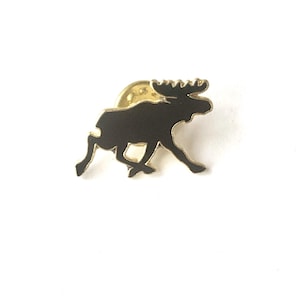 May include: A black moose pin with a gold-colored border and backing. The moose is depicted in a running pose, with its antlers and legs clearly defined. The pin is set against a white background.
