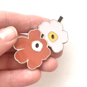 May include: A pair of enamel flower hair clips. One flower is white with a yellow centre and the other is brown with a white centre.