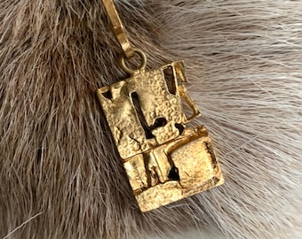 May include: A gold-colored pendant necklace with a rectangular, textured pendant. The pendant has an abstract design with cut-out shapes. The necklace is displayed against a backdrop of fur.