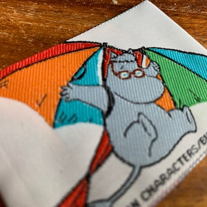 Puede incluir: Un parche de tela blanco con un hipopótamo gris de dibujos animados que lleva gafas y sostiene un paraguas a rayas de colores. El texto "Characters/Bulls/Telec" está impreso en la esquina inferior derecha del parche.