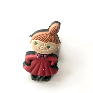 May include: A figurine of Little My from the Moomin series. The figure has a red dress, black shoes, and a bun hairstyle. The character has a stern expression with green eyes and a purple bow tie.