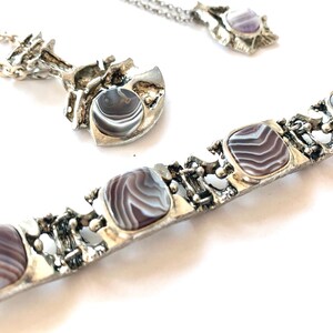 Puede incluir: Un conjunto de joyas plateadas que incluye un collar, un colgante y una pulsera. Las piezas tienen un diseño texturizado y orgánico y presentan piedras cuadradas y redondas con un patrón a rayas, similar al ágata, en tonos grises y blancos. Las joyas se exhiben sobre una superficie blanca.