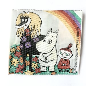 Puede incluir: Etiqueta tejida con personajes de Moomin. La etiqueta muestra una figura negra con cabello rubio, una criatura blanca y una pequeña figura roja, sobre un arcoíris y hojas de otoño. El texto "MOOMIN CHARACTERS/BULL'S" es visible.