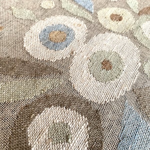 Puede incluir: Primer plano de un textil tejido con un estampado floral. El diseño presenta grandes flores estilizadas en tonos de blanco, azul, verde y marrón sobre un fondo beige neutro. Se aprecia la textura del tejido.