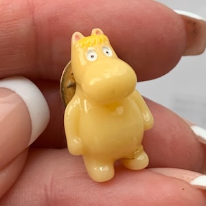 May include: A small, yellow Moomin character pin. The pin features a rounded, cartoonish figure with small arms and legs, and a small gold-colored pin backing. The Moomin has a light yellow body, with a slightly darker yellow hair and small, round eyes.