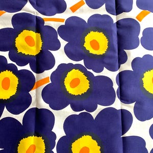May include: A fabric featuring a bold floral design. The pattern showcases large, stylized flowers in deep blue, yellow, and orange against a white backdrop. Each flower has a yellow centre with an orange dot.