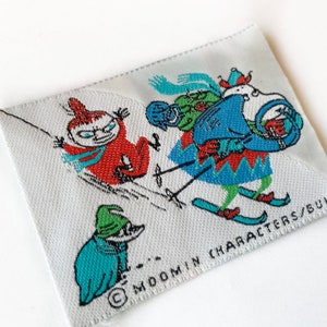 Könnte beinhalten: Ein weißer Stoff-Patch mit einer bunten Illustration von Comicfiguren beim Skifahren. Der Text "MOOMIN CHARACTERS/BULLS" ist unten auf dem Patch aufgedruckt.