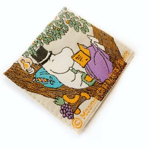 Könnte beinhalten: Ein beigefarbener Stoff-Patch mit einer Cartoon-Illustration eines weißen Flusspferdes, das einen blauen und grünen Hut trägt und ein Buch liest. Das Flusspferd sitzt auf einem braunen Ast mit grünen Blättern und lila Blüten. Der Text "Moomin" ist in Gelb auf dem Patch gedruckt.