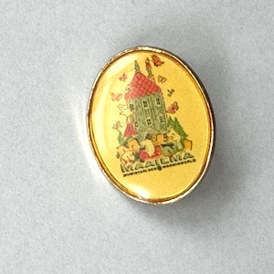 May include: An oval-shaped metal pin with a gold-toned border and a yellow background. The center features a graphic illustration of a tall, multi-story house with a red roof, surrounded by small butterflies. The text MAAILMA MOOMINWORLD is printed.