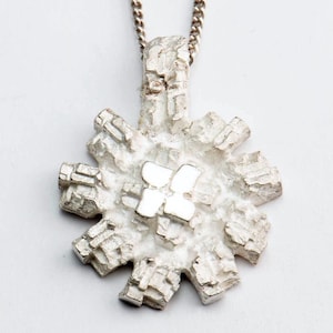 May include: A silver pendant necklace with a snowflake design. The snowflake is made of many small, intricate pieces that create a textured look. The pendant is attached to a silver chain.