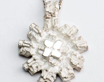 Puede incluir: Un collar con un colgante de plata con forma de copo de nieve. El copo de nieve está hecho de muchas piezas pequeñas e intrincadas que crean un aspecto texturizado. El colgante está unido a una cadena de plata.