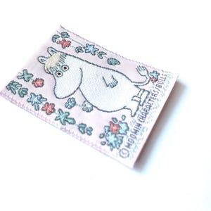 May include: A pink fabric patch with a white Moomin character, with blue eyes and a brown nose, standing in a field of flowers. The text "Moomin Characters Ltd." is printed on the bottom right corner of the patch.