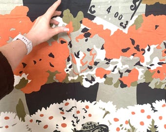 May include: A patterned fabric featuring a design with orange, white, and black elements. The design includes stylized floral and tree-like shapes, with the text "LA BOTTE" and numbers. A hand with a white watch is visible.